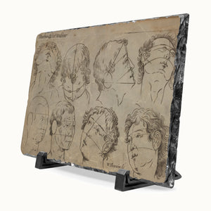 A rectangular stone wall plaque featuring an antique-style sketch of classical facial profile studies on a beige background, mounted on a black marble border and displayed on a black stand against a white background.