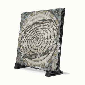 A square decorative plaque or tile with a spiral optical illusion design in beige, gray, black, and muted blue tones, displayed on a black stand against a light off-white background.
