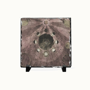 A square decorative slate tile featuring an antique celestial map design with moon phases, sun rays, and astrological symbols, displayed on a small black stand against a light off-white background.