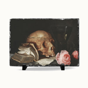 A rectangular slate serving board with a dark still life print of a human skull, an open book, and pink roses, displayed on a white background with two small black stands.