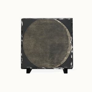 A square black slate art piece featuring a large circular design in textured gray and brown tones, displayed on a small black stand against a plain white background.