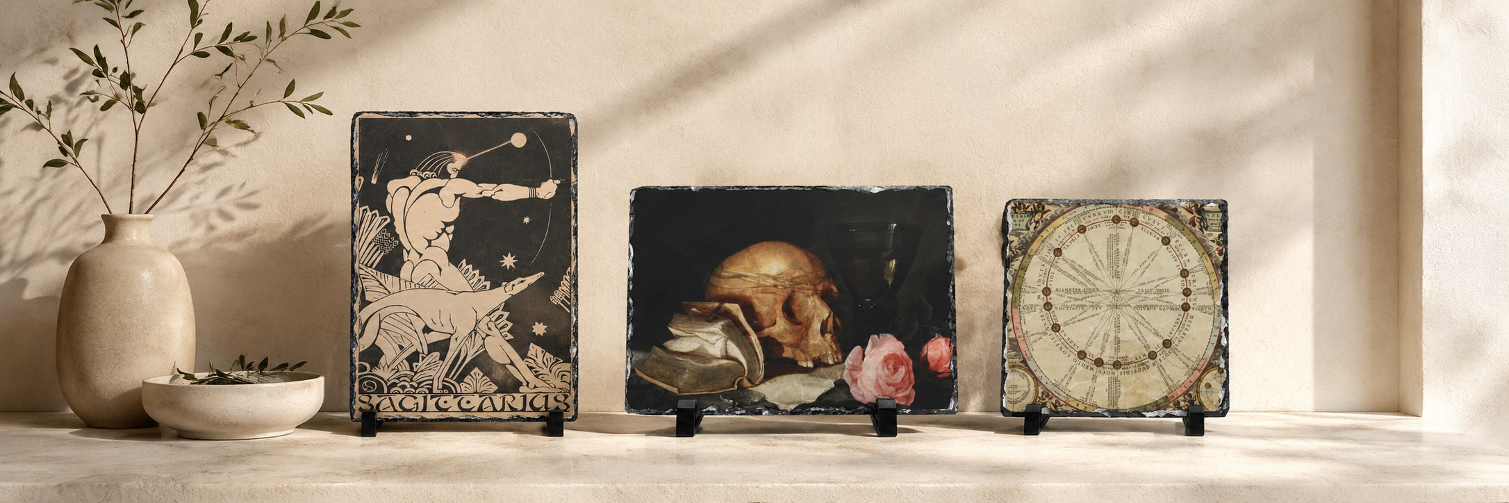 Three square decorative art tiles displayed on a shelf against a sunlit beige wall. The left tile shows a black-and-white classical figure illustration, the center tile depicts a still life with a skull and pink flower on a dark background, and the right tile features an antique-style clock face.