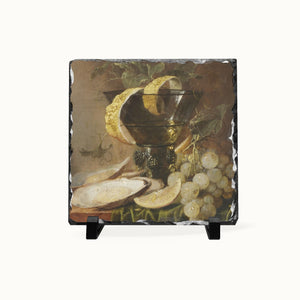 A square slate decorative tile featuring a still life painting of grapes, a lemon, and a bowl, displayed on a small black stand against a light gray background.
