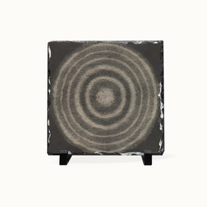 A square black slate coaster featuring a textured beige and gray concentric circle design, displayed on a small black stand against a clean white background.
