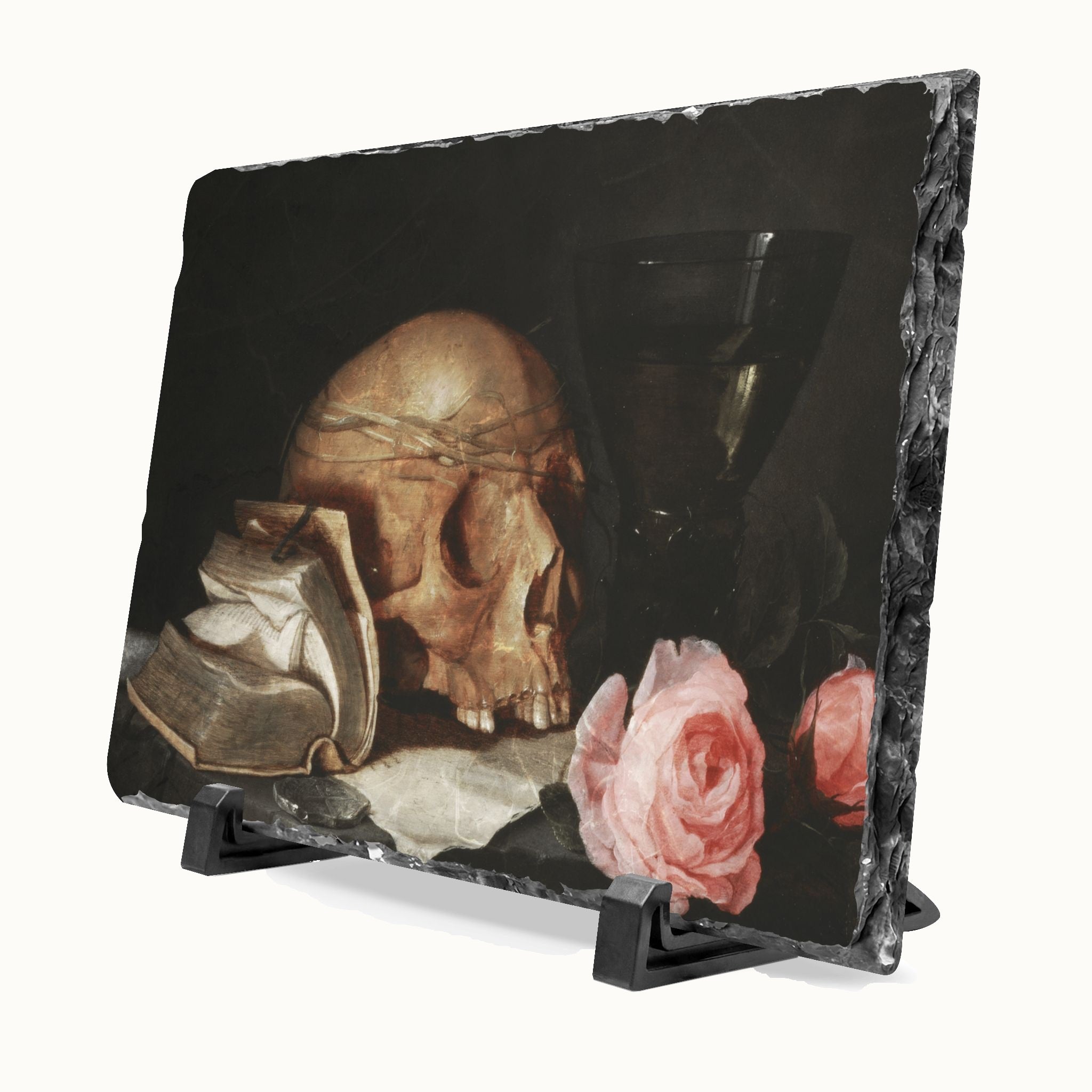 A rectangular black slate plaque with a still life print of a human skull, pink roses, and an open book, displayed on a black stand against a white background.