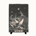 A rectangular black slate art plaque featuring a white line-art illustration of two fish and celestial elements, with the word 'PISCES' at the bottom, displayed on a small black stand against a light background.