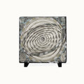 A square decorative tile or coaster featuring a spiral geometric pattern in cream, gray, and black, with a distressed money-print texture and small black display feet at the bottom, shown on a plain light background.