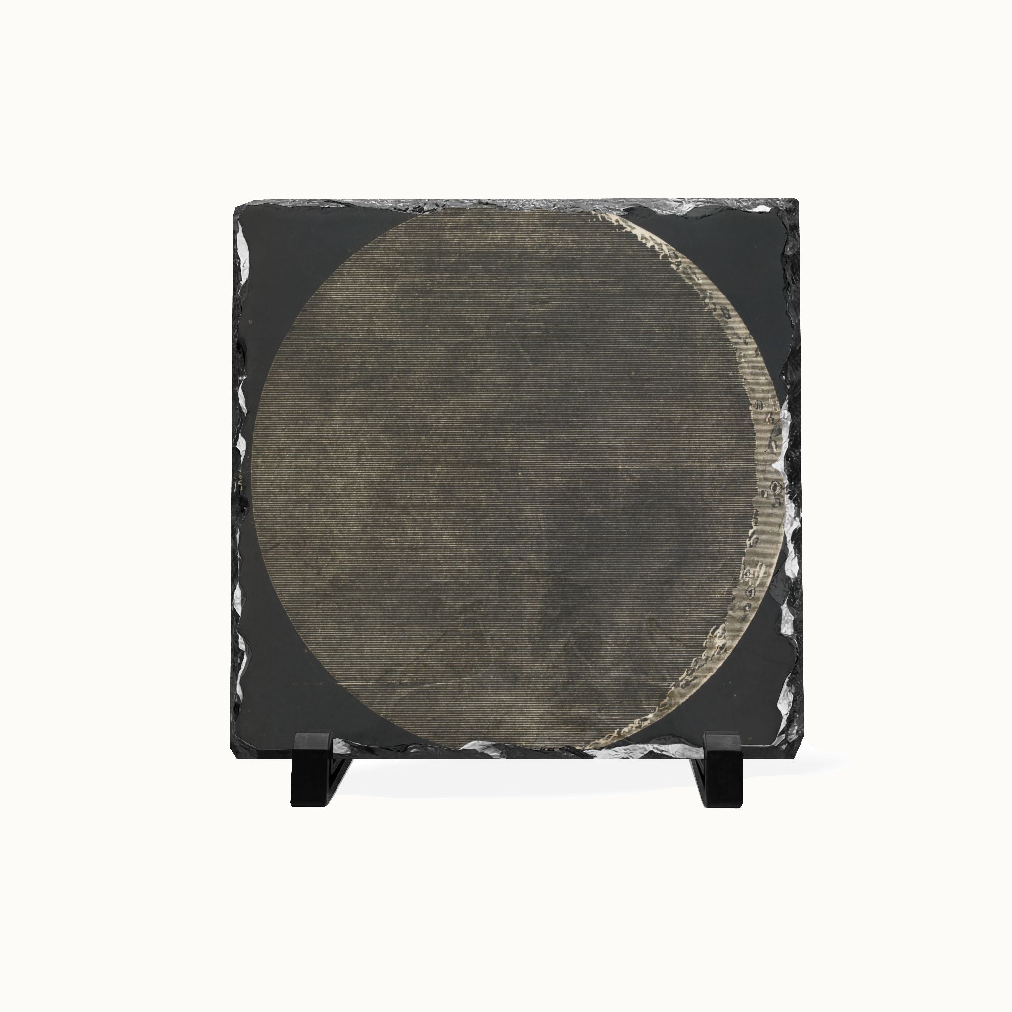 A square black slate art piece featuring a large circular design in textured gray and brown tones, displayed on a small black stand against a plain white background.
