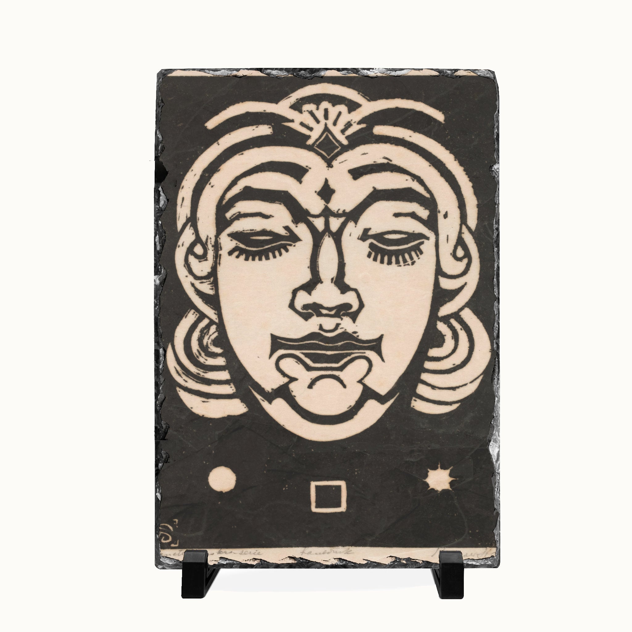 A rectangular black slate art plaque featuring a stylized beige woodcut-style face, displayed on a small black stand against a plain white background.