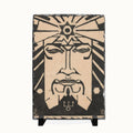 A rectangular decorative plaque or tile featuring a stylized black-and-beige face with geometric star and sunburst motifs, displayed on a small black stand against a plain white background.