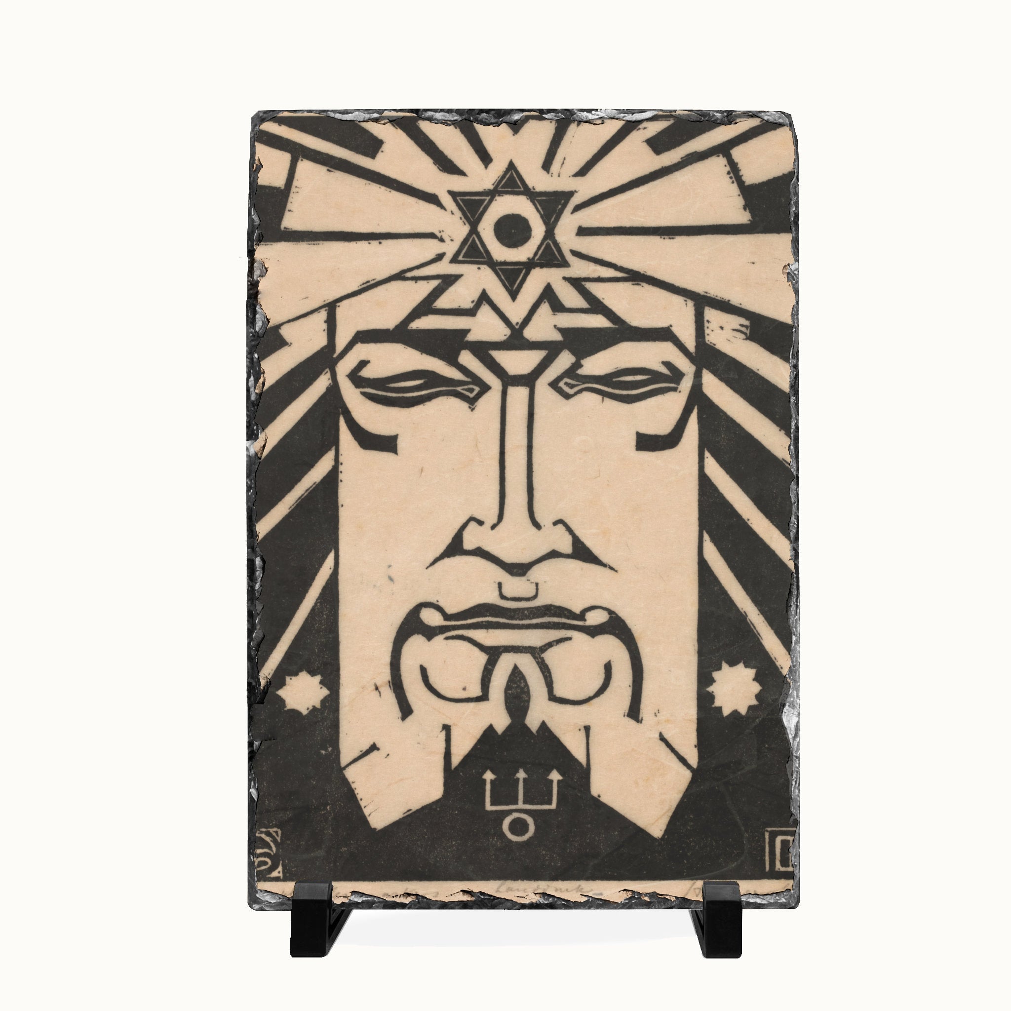 A rectangular decorative plaque or tile featuring a stylized black-and-beige face with geometric star and sunburst motifs, displayed on a small black stand against a plain white background.