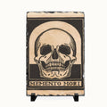 A rectangular slate sign featuring a black and beige skull graphic with the text 'MEMENTO MORI' at the bottom, displayed on a small black stand against a light background.