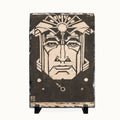A rectangular dark wood plaque or box with a carved face design in light natural wood, displayed on a small black stand against a plain white background.