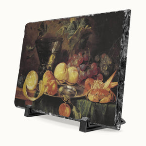 A rectangular black slate serving board with a rustic natural edge, featuring a printed still life painting of fruit, peaches, grapes, and a bowl. The board is displayed on a small black stand against a light gray background.