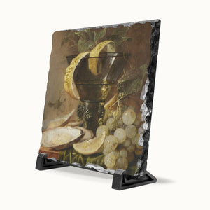 A rectangular slate serving board with a rustic black edge, featuring a still life print of grapes, lemon slices, and a dark goblet. The board is displayed on a black stand against a clean off-white background.