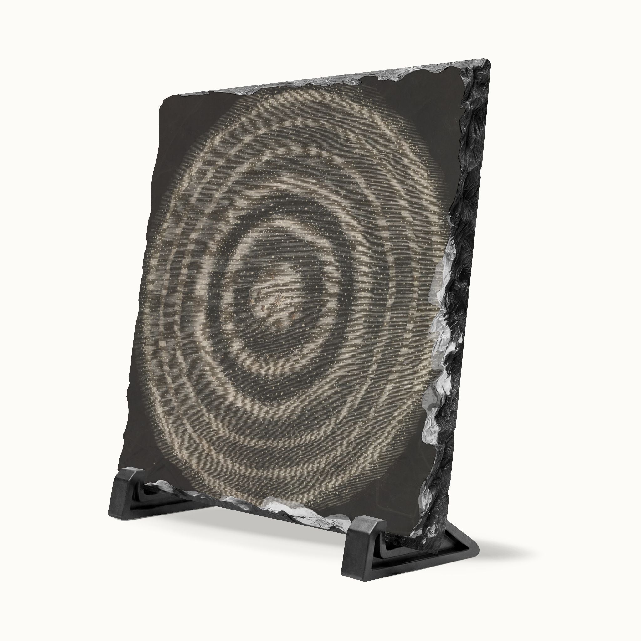 A square black slate art tile featuring concentric circular patterns in shades of gray and beige, displayed on a black stand against a clean off-white background.