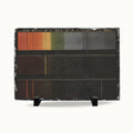 A rectangular black slate art plaque on a small black display stand, featuring a horizontal color gradient from orange and yellow to green and dark gray, photographed against a clean white background.