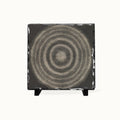 A square black slate coaster featuring a textured beige and gray concentric circle design, displayed on a small black stand against a clean white background.