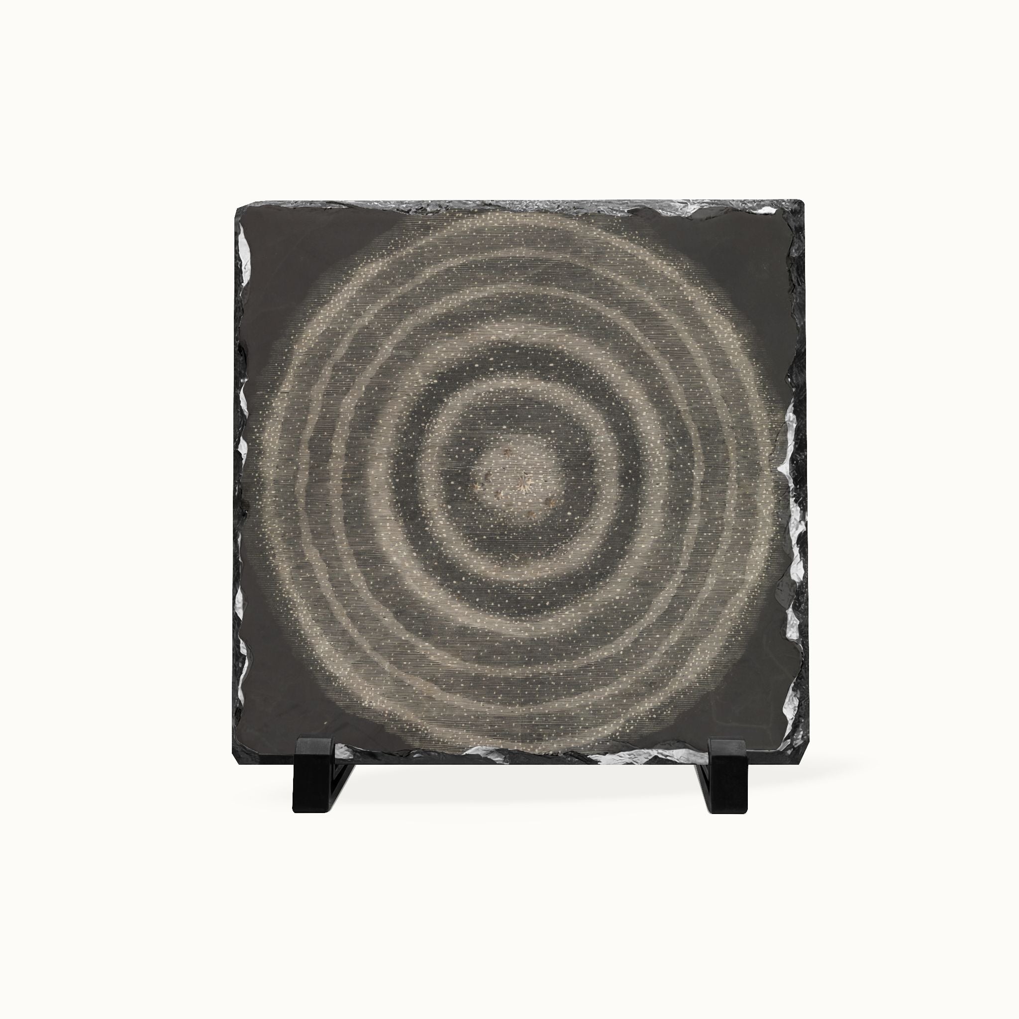 A square black slate coaster featuring a textured beige and gray concentric circle design, displayed on a small black stand against a clean white background.